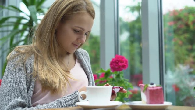 young attractive girl sit in a restaurant in front of a large window. There are flowers on the window sill. attractive blonde takes pictures of food. air curd pink cake on a whipped plate and a cup of