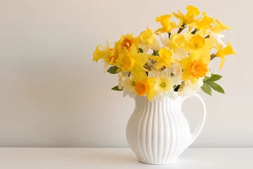 Deurstickers Narcis Close-up van gevarieerde gele narcissen in witte kruik op tafel tegen een neutrale muurachtergrond
