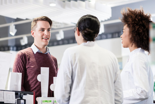 Cheerful young man and happy customer talking with two helpful female pharmacists in a contemporary drugstore with friendly employees