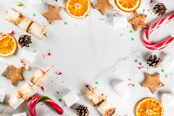 Traditional christmas sweets - candy cane, marshmallow, dried orange, gingerbread stars, baked on fire marshmallow skewers on white marble background, copy space top view