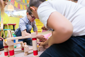 Obraz na płótnie Canvas Cute boy wearing eyeglasses while building with attention and patience a wooden structure during an educational group activity at the kindergarten