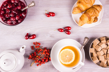 cup of tea with lemon, a teapot, jam from autumn berries and croissants on a rustic wooden background