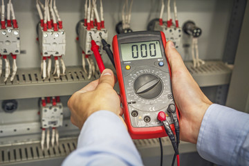 Engineer's hands with multimeter close-up against background of terminal rows of electric automation panel. engineer tests industrial electrical circuits with multimeter in control terminal box
