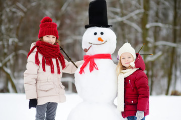 Two adorable little girls building a snowman together in beautiful winter park. Cute sisters...