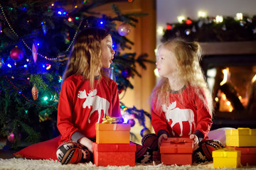 Happy little sisters wearing Christmas pajamas playing by a fireplace in a cozy dark living room on Christmas eve