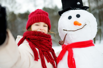 Adorable little girl building a snowman in beautiful winter park. Cute child playing in a snow.
