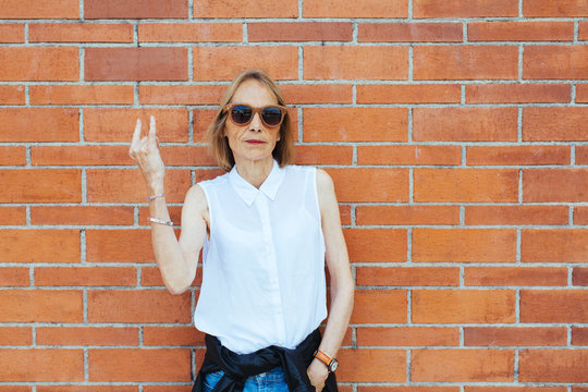 Serious senior woman doing the rock 'n' roll sign in front of a brick wall.