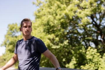 Attractive blonde man wearing a blue shirt, standing leaning back outside on a sunny summer day looking away from camera.