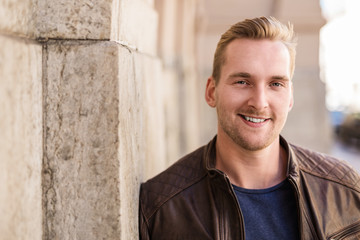 Portrait of a blonde man wearing a brown leather jacket leaning against a stone wall outdoors on a sunny summer day.