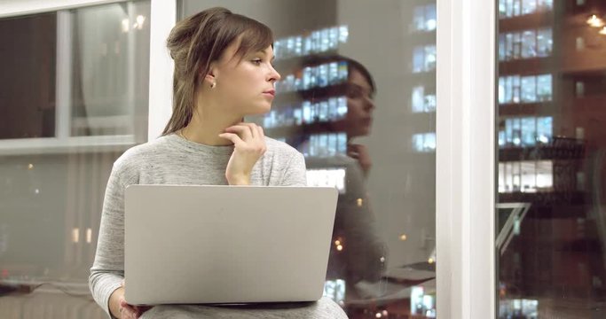 Young woman at home flat working laptop computer and looking window 4k video. Girl in apartment with night city landscape typing on keyboard