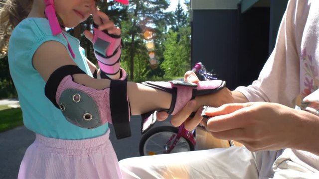 Careful mother helps her little daughter to wear protective elbow pads and gloves