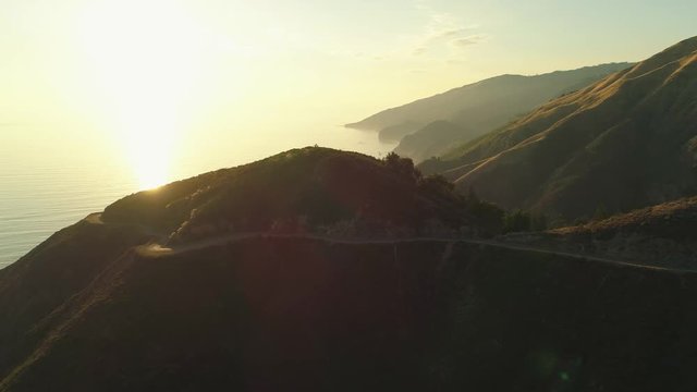 Aerial shot revolves around a hilltop overlooking the ocean with a windy road. Sunset reflects off water.