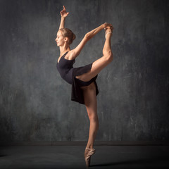Fototapeta premium A young beautiful ballerina in a black dress, white pantyhose and pointe shoes beautifully poses and dances ballet in an image in a dark dance stage