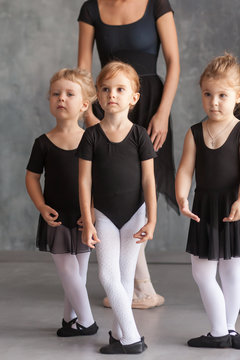 a young woman ballerina in a black dress, white pantyhose and pointe shoes teaches to dance the ballet of young girls ballerinas in black dresses and tights in a dark dance studio