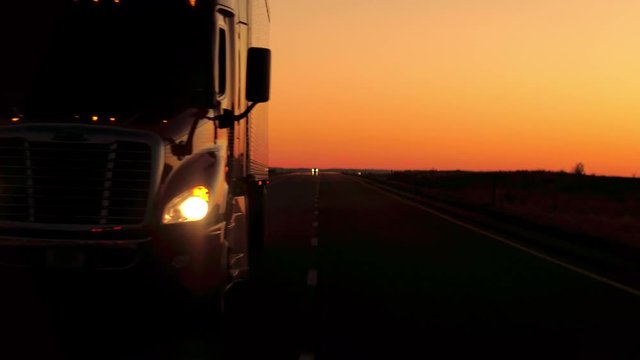 CLOSE UP: Front view of freight container semi trailer truck transporting goods driving along multiple lane country highway in Great Plains after the sunset. Overtaking lorry traveling across the USA