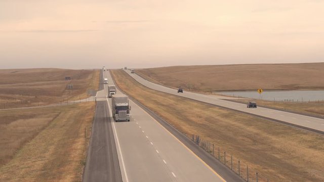 AERIAL, CLOSE UP: Freight container semi truck hauling goods, cars, SUVs and pickups traveling along the Great Plains highway running through vast prairie fields in wild American countryside at dusk