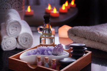 Obraz na płótnie Canvas Set for spa procedures - stones for massage, oil, sea salt of violet and natural amethyst lies in a box. Moist towels lie on a natural black granite table. Electric candles burn in the background.