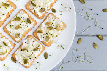 Tasty crackers with cream cheese, seeds and greens. Appetizers on a plate on grey table. Healthy snacks, top view, flat lay