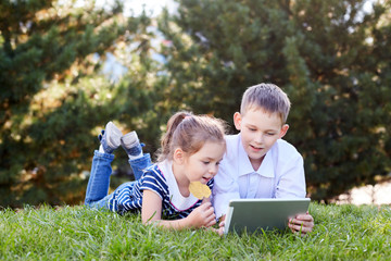Brother and sister lying on the grass in the park and play tablet. Children watch cartoons on the tablet.