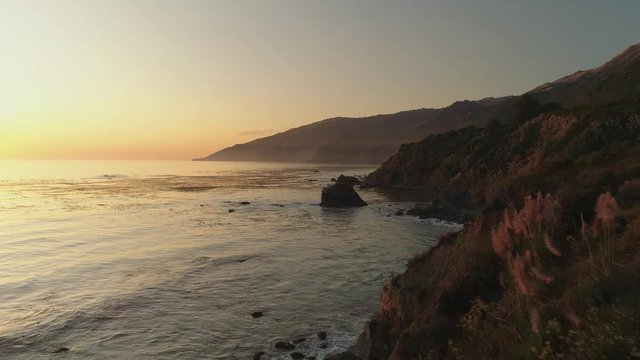 Aerial shot approaching ocean waves at sunset by a rocky coast. Orange glow, clear skies.