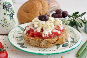 Dakos traditional greek appetizer on a traditional plate with ceramic olive oil jar, dry rye bread, olives and olive branch. Healthy eating concept. Mediterranian lifestyle - obrazy, fototapety, plakaty