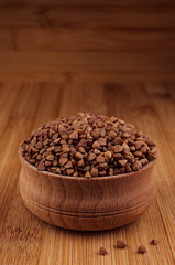 Fototapeta na wymiar Buckwheat brown in wooden bowl on brown bamboo board, closeup. Rustic style, healthy dietary groats background.