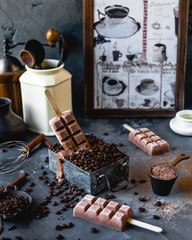 coffee popsicles on wooden sticks with melting popsicle in metal box filled with coffee beans, with cinnamon, cocoa powder, carnations, coffee pot and grinder on gray background