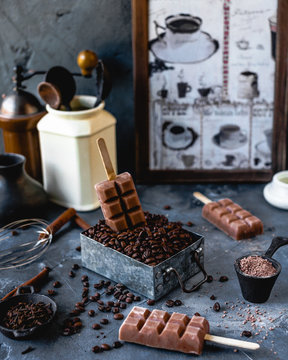 Coffee Popsicles On Wooden Sticks With Melting Popsicle In Metal Box Filled With Coffee Beans, With Cinnamon, Cocoa Powder, Carnations, Coffee Pot And Grinder On Gray Background