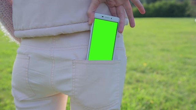 Young Woman staying in city park pulls white SmartPhone with pre-keyed green screen out of her pocket. Perfect for screen compositing. Made from 14bit RAW. 10bit ProRes 444