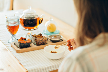 Close up of woman's hands holding spoon and bowl with granola - healthy morning breakfast concept. Breakfast table with granola, yoghurt, tea, lemon and bowls with almond, dried apricots, jam, kettle.