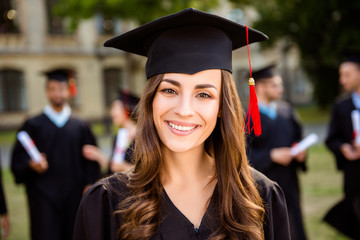 Happy cute brunette caucasian grad girl is smiling, her class mates are behind. She is in a black...