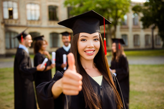 Close up portrait of attractive asian student showing agree sign. She is in gown, moratr board, with red tassel, behind her are her classmates, in a green park near the building of campus