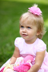 Sweet smiling little girl with long blond hair, sitting on grass in summer park, closeup outdoor portrait. Adorable little girl on grass. Little girl in the garden 