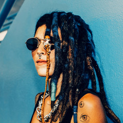 Rasta Spanish Girl with dreadlocks, piercings, tattoos and stylish accessories on the blue wall.