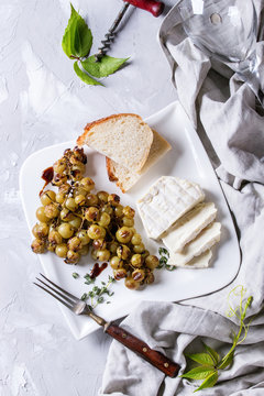 White plate with sliced camembert cheese and baked bunch of green grapes served with bread, wine glass, corkscrew, green leaves, fork on textile over gray texture background. Top view with space