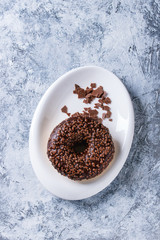 Chocolate glazed donut with chopped chocolate on white plate over gray texture background. Top view with space