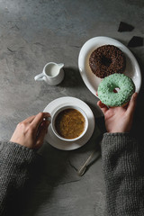 Plate of colorful glazed donuts with chopped chocolate, cup of black coffee, jug of milk over gray texture table. Female hands taking turquoise donat. Flat lay with space