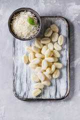 Raw uncooked potato gnocchi on white wooden chopping board with ingredients. Flour, grated parmesan cheese, basil over gray concrete background. Top view with space. Home cooking.
