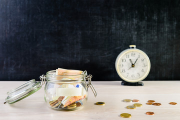 Fototapeta na wymiar Coins and paper money in a glass jar on wooden table against dark background. Behind table clock, concept of investment, savings and business growth