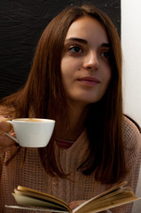 A young beautiful girl in a warm sweater holds a cup of coffee and reads a book, a cute portrait