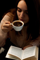 A young beautiful girl in a warm sweater drinks coffee and reads a book, a cute portrait