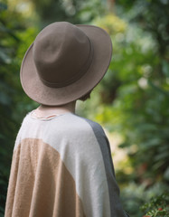 Fashionable young woman wearing in hat and poncho among tropical plants. Rear view.