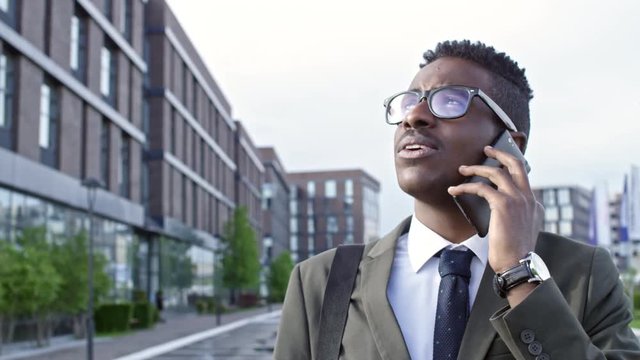 Slowmo of African businessman in eyeglasses standing outdoors in downtown and talking on the phone