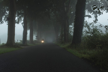 Misty country road with scooter and illuminating headlight.