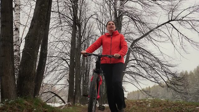 The woman costs with bicycle of a pn a background of the avenue from trees. The woman in an orange jacket walks with bicycle. The hostess walks the dog. Avenue in the countryside. Nature.
