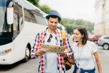 Cheerful guy in hat and shirt, holding city map, looking with smile in it, standing near his female...