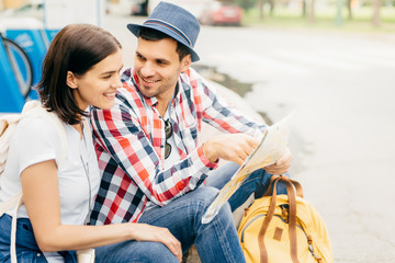 Portrait of happy male and female friends, having trip, resting on park bench, looking happily into map, choosing place where to go. Young tourists having vacation using city map. Relaxation concept