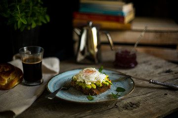 Black bread toast with fried benedict egg, avocado and salmon spread in blue plate with coffee in background, wooden shabby table with coffee pot, glass and books stack
