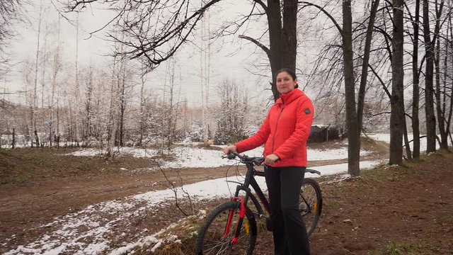 Active girl cycling with her dog. The woman costs with bicycle of a pn a background of the avenue from trees. The woman in an orange jacket walks with bicycle. The hostess walks the dog. Avenue in the
