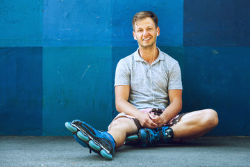 Happy handsome young roller in inline skates sitting against blue wall.